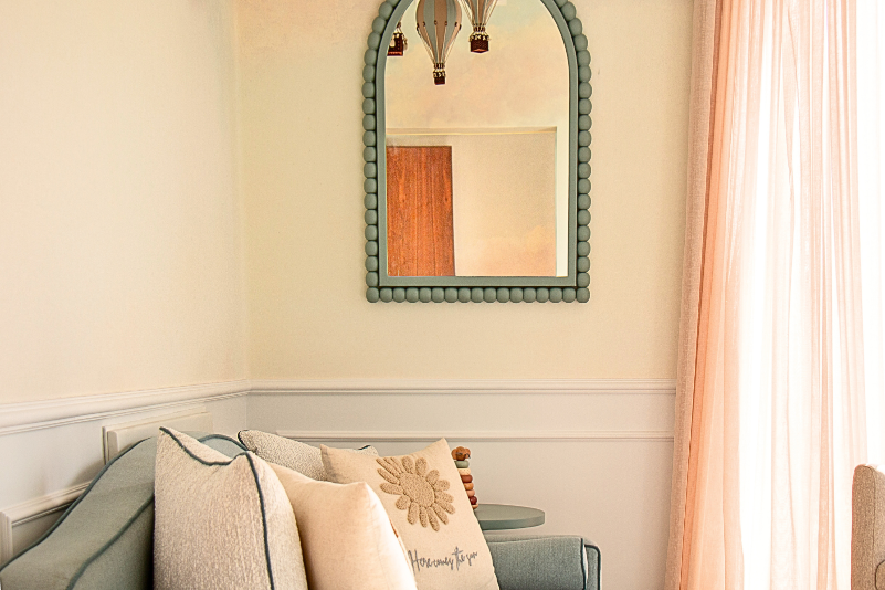 Miroir décoratif au cadre arrondi au-dessus d'une banquette dans une chambre de bébé douce et lumineuse, avec moulures murales Orac blanches. Design mural élégant et apaisant.