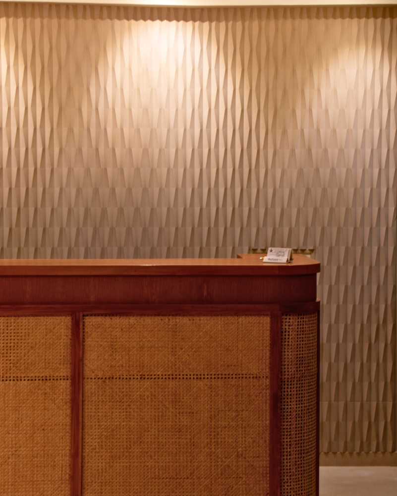 Mur d’accueil décoré avec le panneau 3D Orac W112 Ridge, éclairé par un jeu de lumières indirectes, créant un relief subtil derrière un comptoir en bois et cannage naturel.