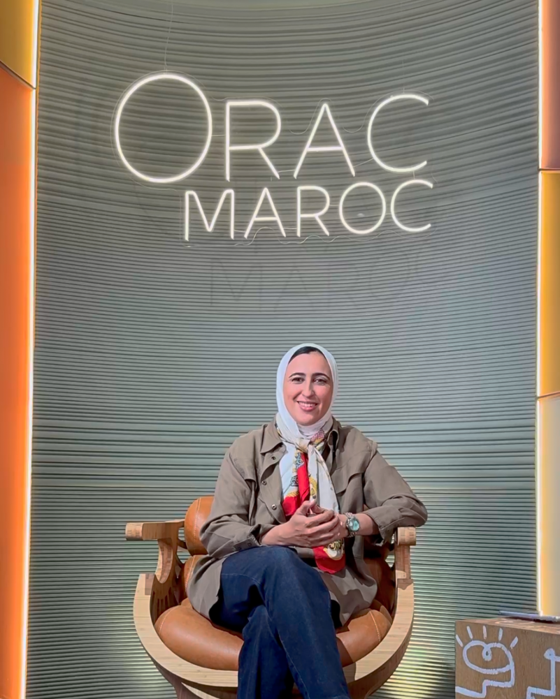 L’architecte Sarah Khalil assise dans le showroom Orac Maroc à Casablanca, devant un mur habillé de profilés décoratifs éclairés. Elle sourit face caméra, évoquant son projet de cabinet médical et son expérience de collaboration avec Orac Maroc.