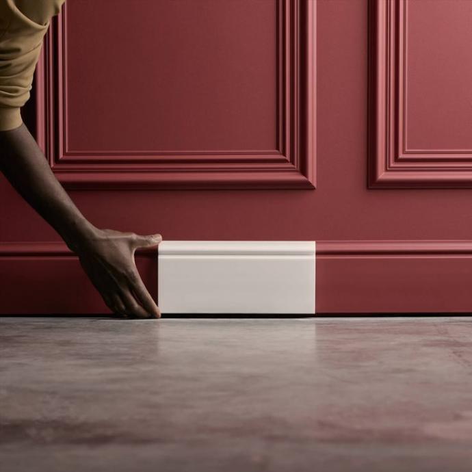 Une main place une plinthe Orac Decor blanche le long d’un mur rouge aux moulures raffinées. La finition nette de la plinthe crée un contraste marqué avec le mur coloré et le sol brut, illustrant la précision et la qualité des profilés Orac.