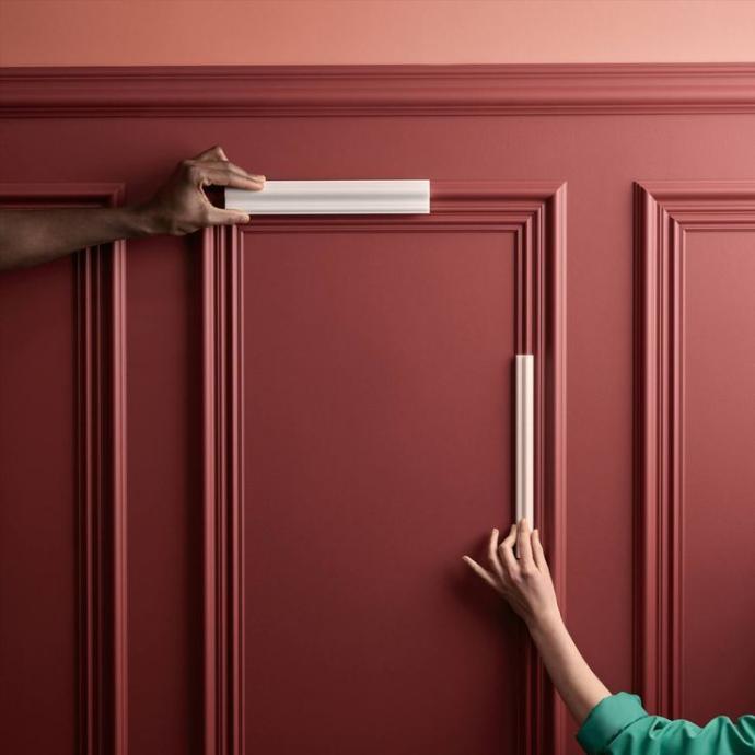 Deux personnes positionnent des profilés Orac Decor blancs sur un mur rouge aux moulures détaillées. L’image met en avant la précision d’ajustement et la qualité des finitions des éléments décoratifs Orac, conçus pour créer des compositions murales harmonieuses.