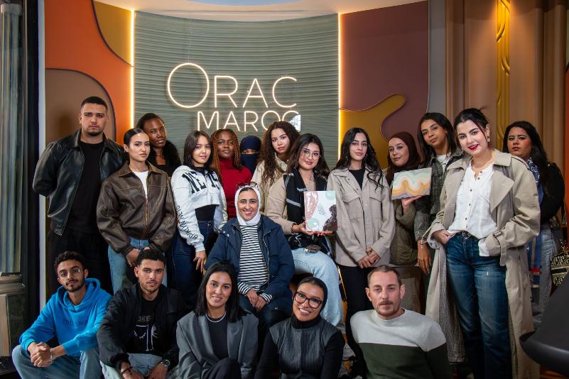 Photo de groupe de participants (étudiants et professionnels) posant devant un mur de marque éclairé 'ORAC MAROC' à la fin d'une Masterclass ou d'un workshop.
